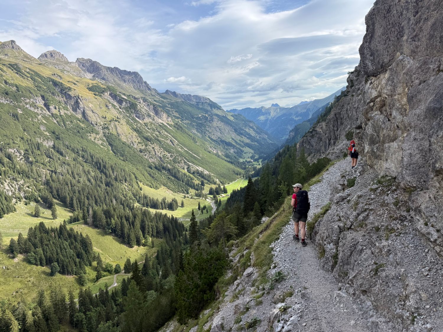 Wanderin mit Rucksack auf einem schmalen Bergpfad an einer Felswand, vor ihr ein weites grünes Alpental