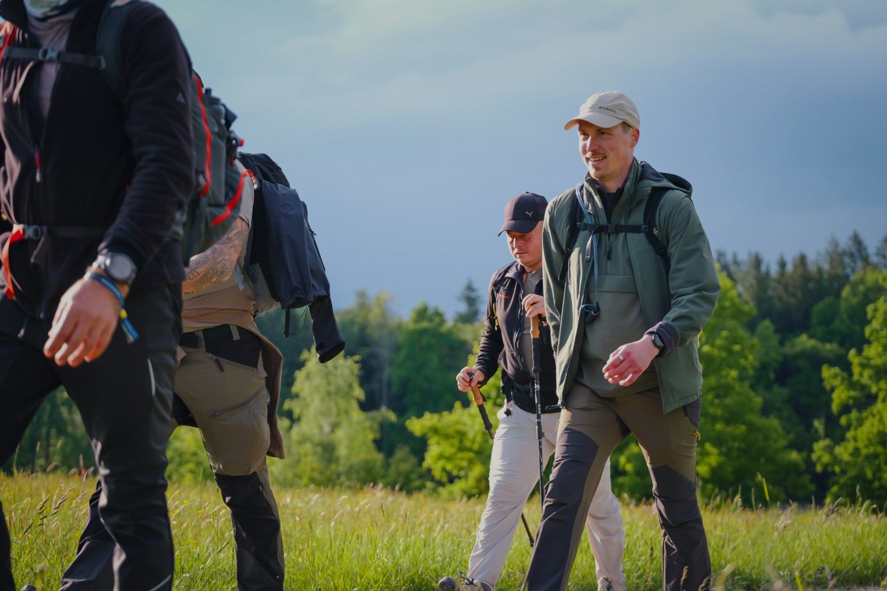 Lachende Wandergruppe mit Rucksäcken auf einer sonnigen Wiese vor dem Waldrand