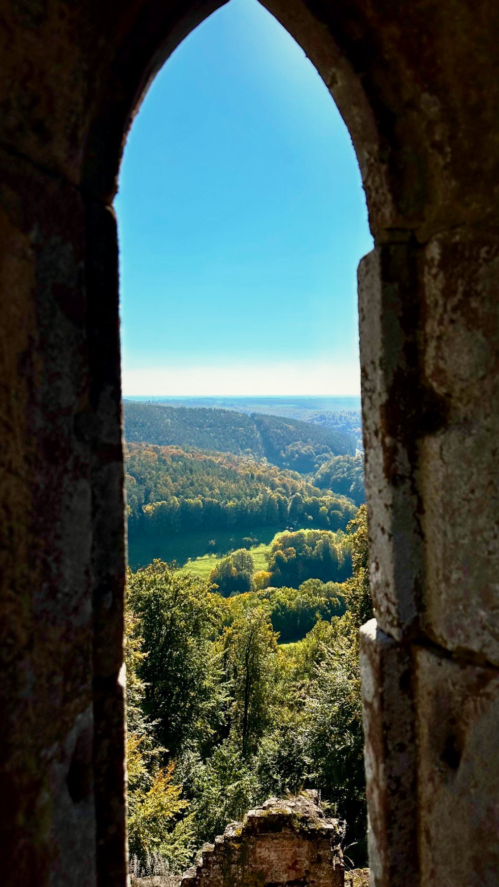Blick durch einen steinernen Torbogen auf ein bewaldetes Tal unter blauem Himmel